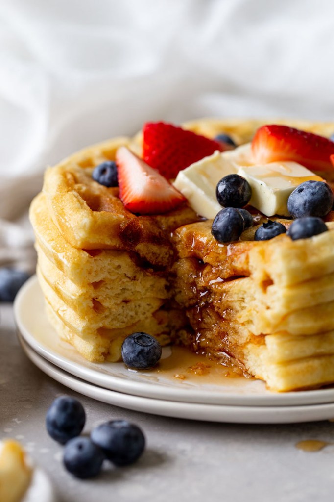 A stack of four fluffy waffles from scratch, topped with fresh fruit, butter, and syrup. A big bite is missing from the stack. 