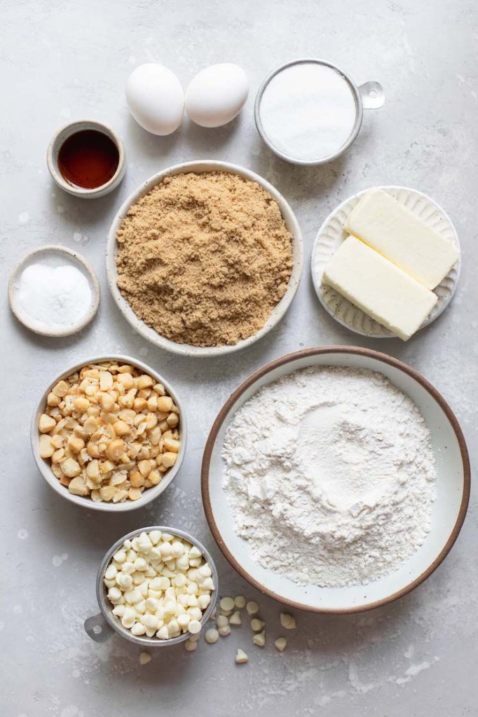 The ingredients needed to make white chocolate macadamia nut cookies in bowls on top of a gray surface.