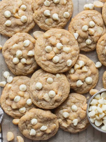 A batch of cookies laid out on a piece of parchment paper with extra nuts and white chocolate chips beside them.