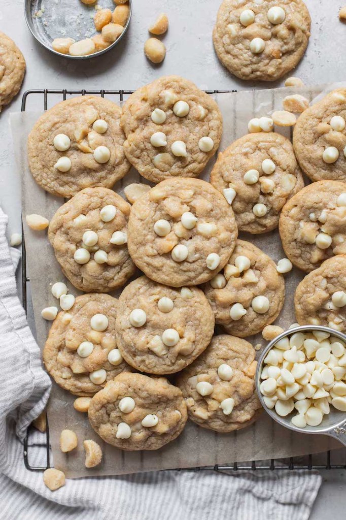 A batch of cookies laid out on a piece of parchment paper with extra nuts and white chocolate chips beside them.