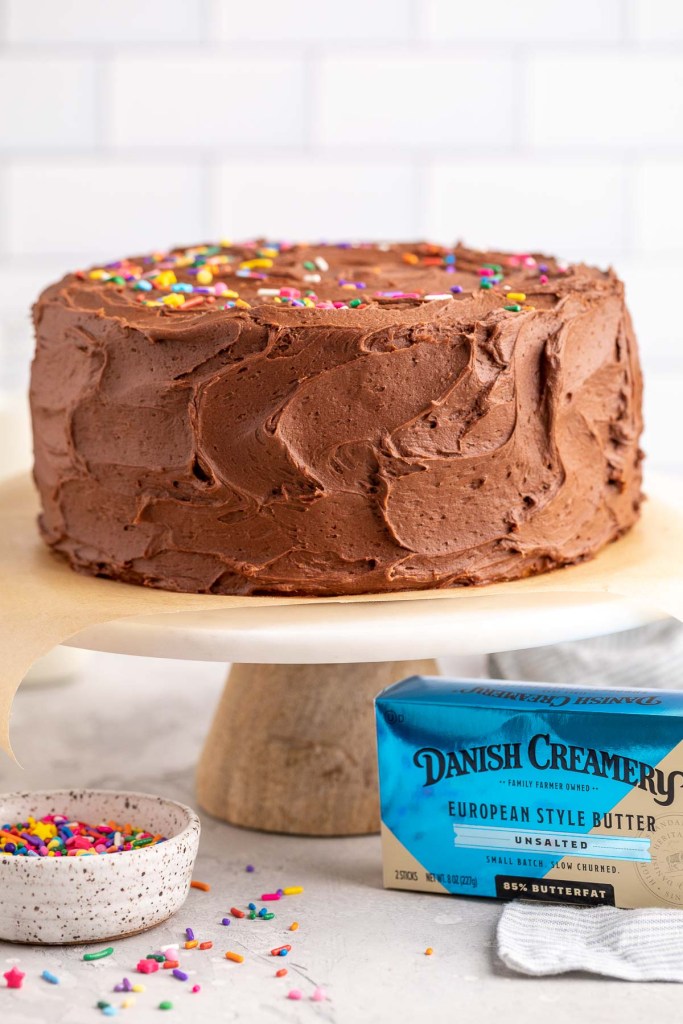 A yellow cake sitting on top of a marble cake stand. A bowl of sprinkles and a box of Danish Creamery butter are sitting next to the cake stand.