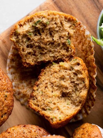 An overhead view of zucchini muffins on a wood cutting board. One muffin has been cut in half and others are lying on their sides.